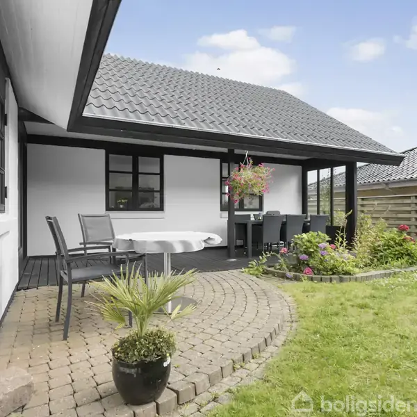 En overdækket terrasse med havemøbler ligger ved siden af et hvidt hus. Terrassen har belægningssten og er omgivet af en velplejet græsplæne og blomsterbed.