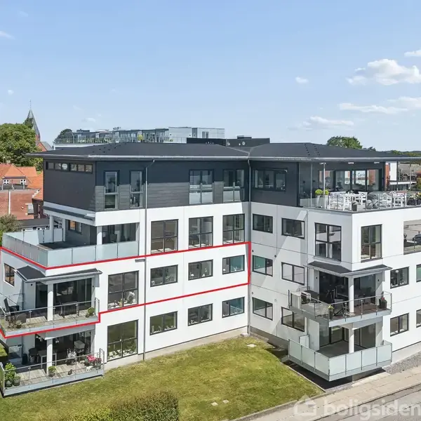 En moderne, fleretagers bygning med altaner. Bygningen er omgivet af en grøn græsplæne og en parkeringsplads. I baggrunden ses en by med huse og biler i solskin.