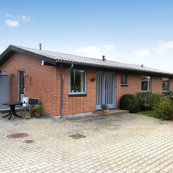 En rød murstensvilla med grå tag og ruder placeret foran en flisebelagt indkørsel, omgivet af buske og en sort bordgruppe nær hoveddøren.