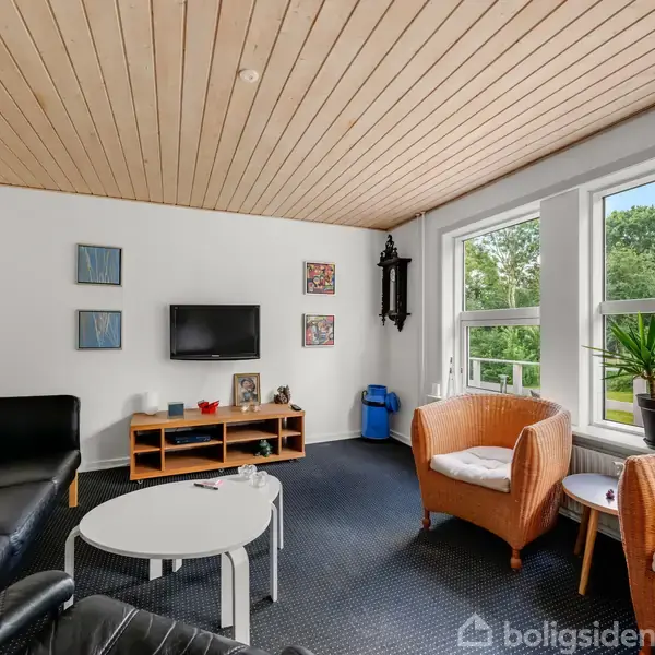 En stue med sorte lædersofaer og kurvestole omkranser et rundt sofabord. Et TV hænger på væggen over en træreol. Store vinduer afslører en frodig have.