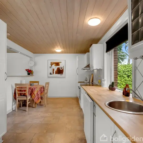 Køkken med træloft har bordplade med vask og køkkenredskaber, set mod spiseområde med træbord og stoler dækket med farverigt tæppe. Store vinduer viser grønne buske udenfor.