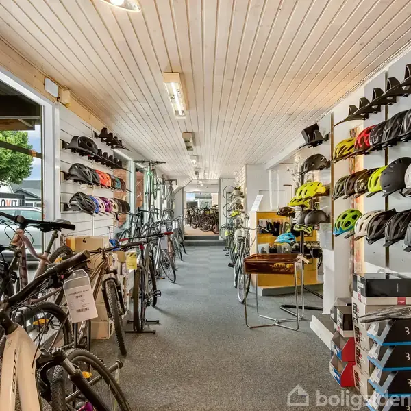 Cykler står på rad langs væggene, mens farverige hjelme er udstillet på hylder i en cykelbutik. Andre cykeltilbehør kan ses i forretningen. Udenfor er en parkeret bil.