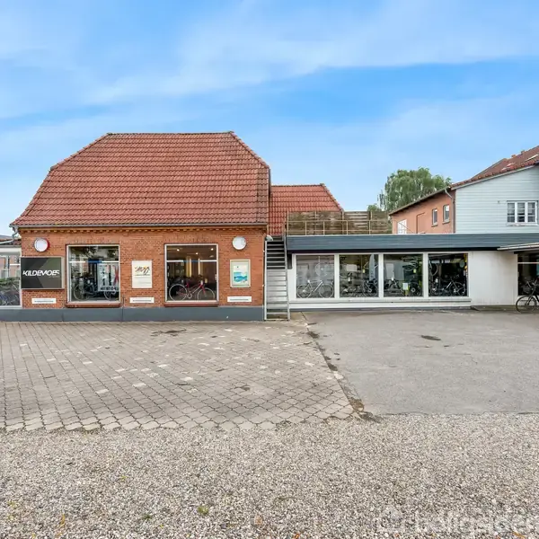 En gulstensbygning med skiltet "MEINECKE" viser cykler i vinduerne. Der er en stor parkeringsplads foran, og bygningen er omgivet af andre strukturer på en solrig dag.