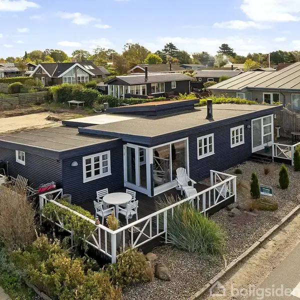 En sortmalet bungalow står stille i et boligområde, omgivet af en velholdt have og en hvid terrasse med havemøbler, på en vej med flere lignende huse i nærheden.