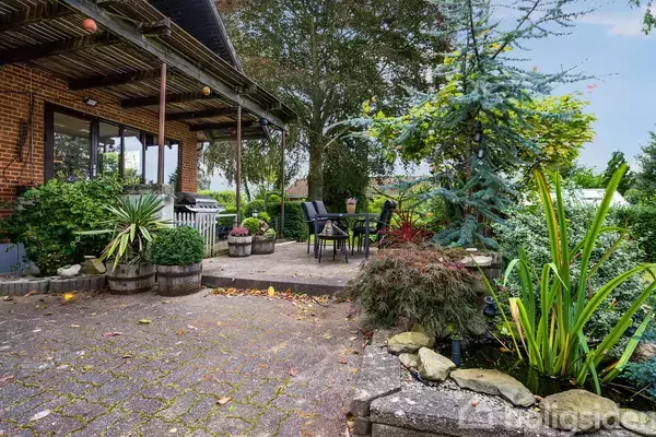 Terrassemøbler står på en flisebelagt terrasse omringet af grønne planter og træer. En træhytte med beskedent tag er synlig i baggrunden, hvilket skaber en hyggelig havelignende atmosfære.