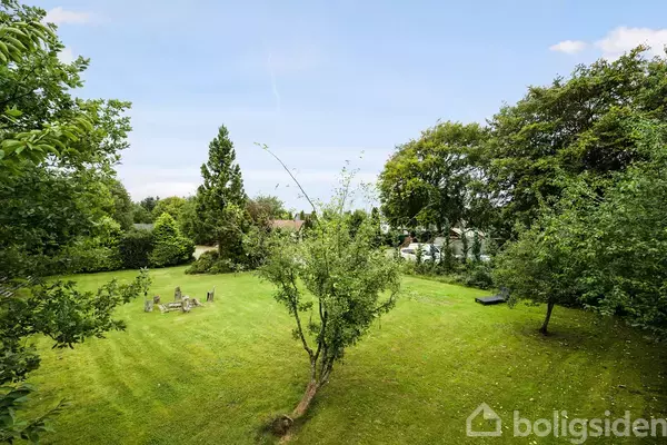 En velplejet græsplæne med et centralt placeret lille træ. Omringet af tætte buske og træer i en stille have. I baggrunden ses en lille cirkel af havemøbler.