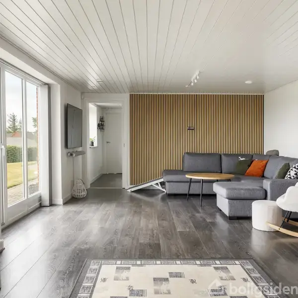 En moderne stue med en grå hjørnesofa og en gyngestol i højre side. En væg med træpaneler skaber en stilfuld baggrund. Store vinduer lukker lys ind.