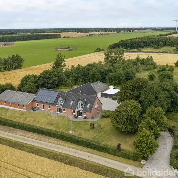 Stor gård med solpaneler på taget omgivet af frodig vegetation og marklandskab. En grusvej løber langs ejendommen, der er omkranset af marker og forskellige træer.