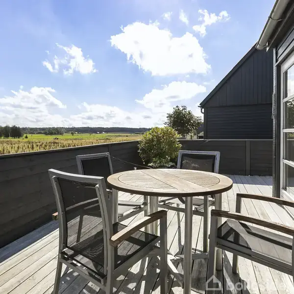 Rundt bord med fire stole står stille på en solrig terrasse af træ. Udsigt over et grønt landskab med marker og spredte træer under en blå himmel med hvide skyer.