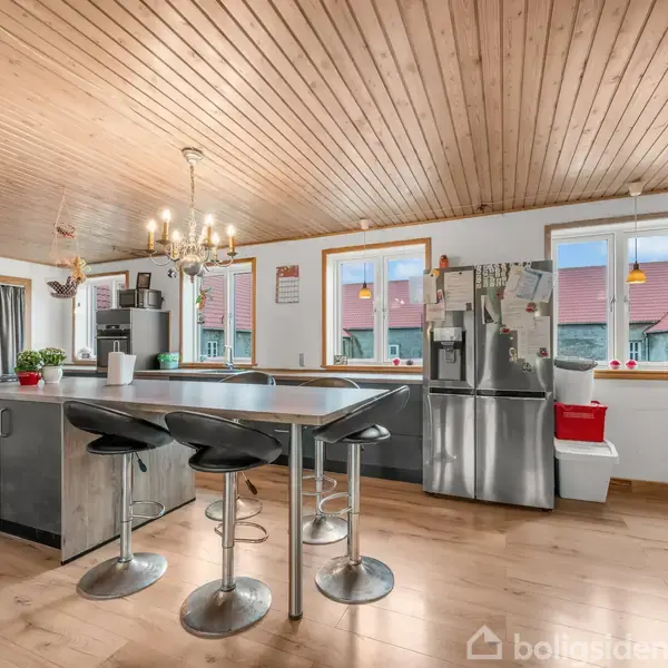 Køkken med køkkenø, tre barstole, og et køleskab. Træbeklædt loft og gulv. Væggene har vinduer med udsigt til tage. Lysekrone hænger over øen.