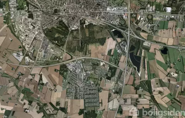 Satellitfoto viser en by med veje og motorveje i et netværk, omgivet af marker og skove. Bygninger er tæt koncentreret i den øverste halvdel af billedet.