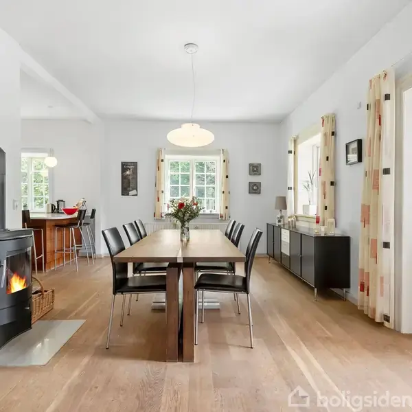 Et spisebord omgivet af stole står i et lyst rum med trægulv. En tændt brændeovn er ved siden af bordet, mens vinduer og en glasdør tillader naturligt lys at strømme ind.