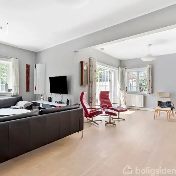 En stor stue med en sort hjørnesofa og vægmonteret TV, flankeret af røde stole. Sollys strømmer ind gennem store vinduer, der afslører en lys træoverflade.