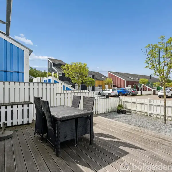 Udendørs terrasse med et sort havebord og fire stole. Sollys skinner på trædækket ved siden af en lav, hvid stakit. Omkring er der parkerede biler, farverige huse og træer.