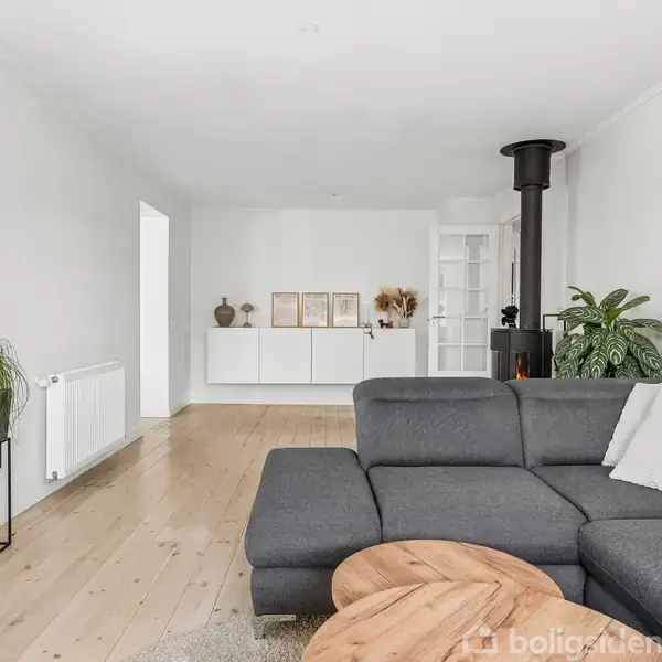 Grå sofa står i stue med træplankegulv, ved siden af en rund træbord. I hjørnet er en sort brændeovn og en stor grøn plante. Væggen har opbevaringshylder.