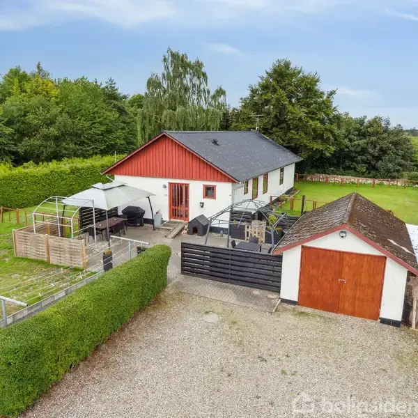 Et enkelt hus med rødt gavl og sort tag, omgivet af en grøn have. På terrassen står havemøbler under en markise. Gårdspladsen har en garage med dør i brun træ.