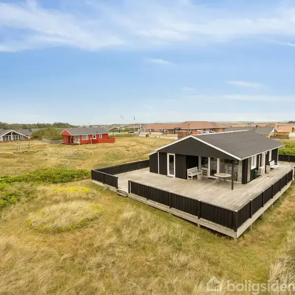Et sommerhus med sort tag og terrasse omgivet af sort stakit står på en græsbeklædt grund i et åbent landskab med andre huse i baggrunden under en blå himmel.