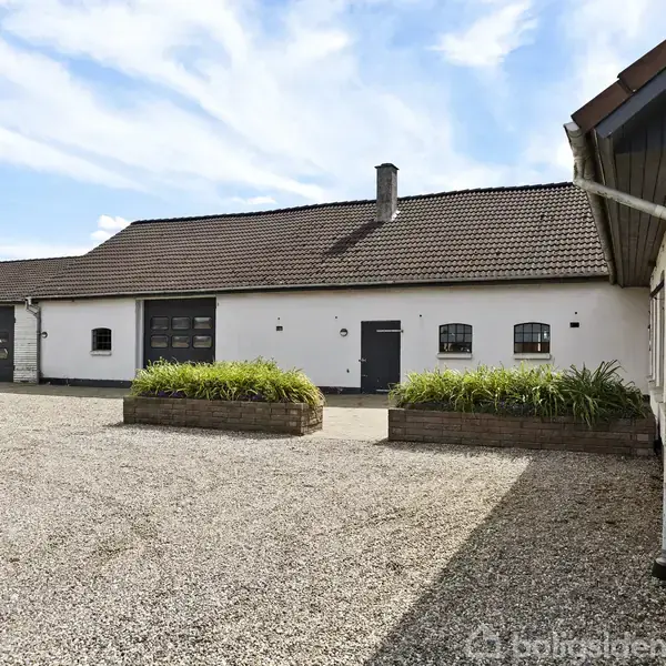 Et hvidt hus med rødt tegltag står i en grusbelagt gårdsplads. Huset har flere små vinduer og en sort dør. Tallet “57” er tydeligt på facaden.