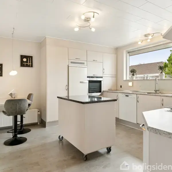 Et moderne køkken har en fritstående ø, hvide skabe og apparater. En stor vindue giver udsigt til et træ udenfor. Spisebord med to stole placeret i hjørnet.