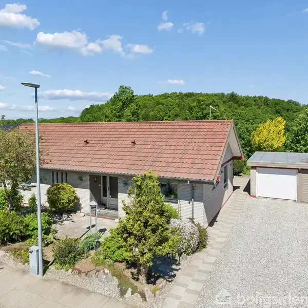 Rødtagset hus med carport ses fra gaden. Grusbelagt indkørsel og velplejet have omkranser huset. Blå himmel og træer i baggrunden.