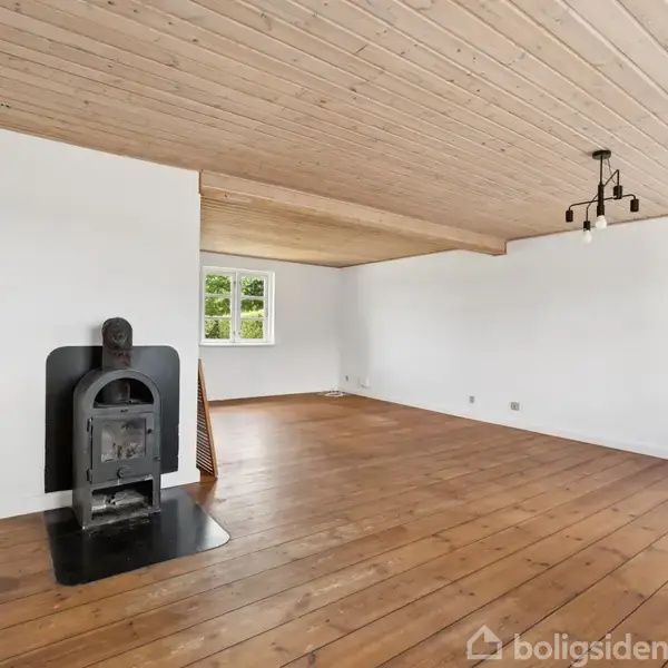 En brændeovn står i hjørnet af en stor, tom stue med trægulv og træloft. Der er et vindue mod væggen, og et enkelt sort loftlys hænger fra loftet.