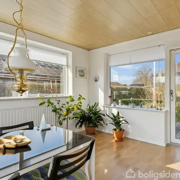 Bord med glasplade står i en lys spisestue, omgivet af planter og en klassisk lampe. Gennem vinduerne og en glasdør ses en terrasse og have i baggrunden.