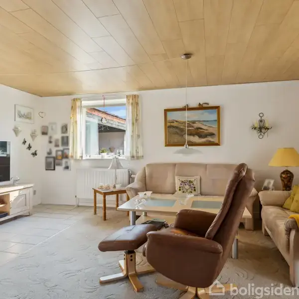 Et hyggeligt stueområde indeholder en brun lænestol og beige sofaer omkring et sofabord; dekoreret med billeder på væggene og en stor TV-skærm til venstre, med dagslys indstrømmende fra et vindue.