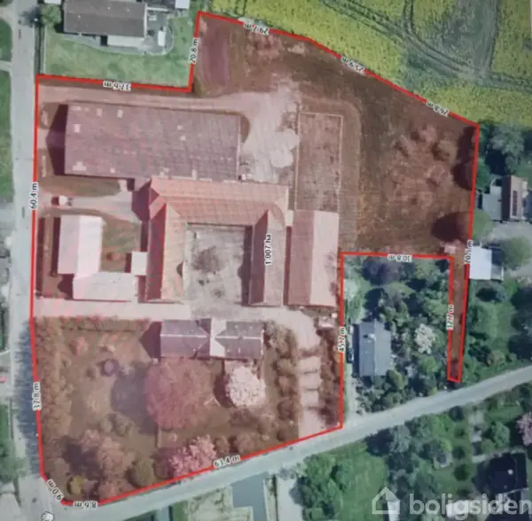Luftfoto viser en landbrugsejendom omgivet af røde linjer angivende grænser. Ejendommen består af flere bygninger med omgivende jordarealer, beliggende nær en vej med parcelhuse.