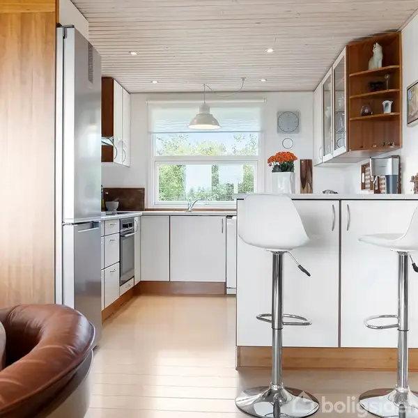 Et moderne køkken med hvide skabe og træelementer, to hvide barstole ved en køkkenø. Store vinduer bringer lys ind i rummet, og en blomstervase står på bordet.