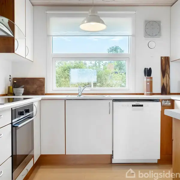 Et moderne køkken med hvide skabe og trækanter. Ovnen og opvaskemaskinen er indbygget. En stor vindue slipper naturligt lys ind og afslører en grøn udsigt udenfor.