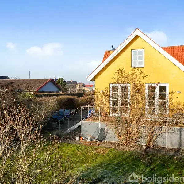 Et gult hus med rødt tag står i en have omgivet af buske og træer. I baggrunden ses flere huse og en blå himmel.