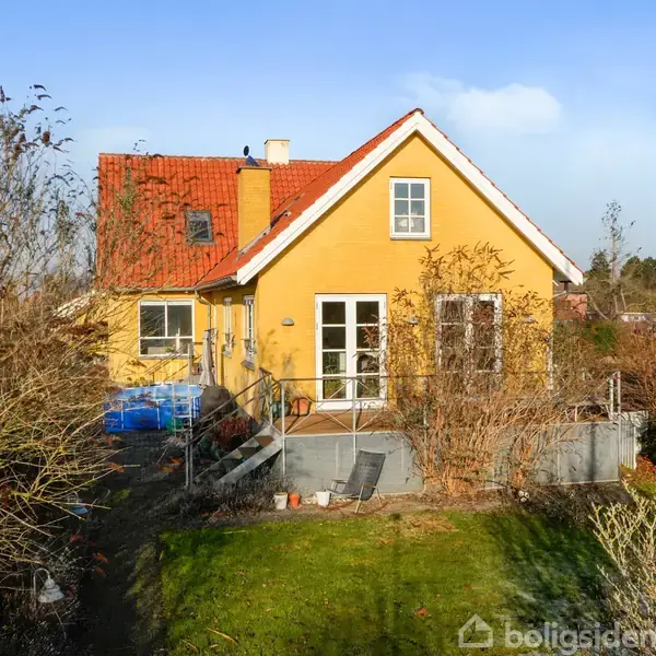 Et gult hus med rødt tag står stille i en grøn have omkranset af buske og træer under en blå himmel.