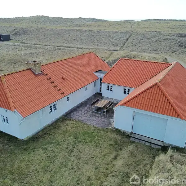 Et hvidt hus med rødt tegltag står på en græsplæne omgivet af klitter. I midten af bygningen er der en gårdsplads med et bord og bænke.