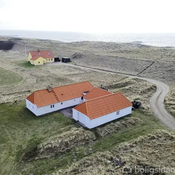 Hus med rødt tag ligger på en græsmark ved kysten. En grusvej fører til et andet gult hus i nærheden. Havet kan ses i baggrunden.