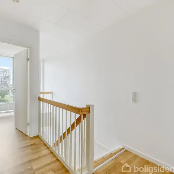 En lys gang med trægulv fører til et åbent rum med en dør. På væggen ses en stikkontakt. Udsigt til en balkon og bygninger udenfor.