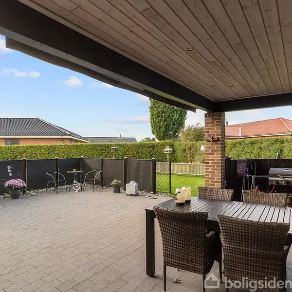 Udendørs terrasse med flisebelægning, rattan bord med stole. I baggrunden står blomsterkrukker og to stole foran en sort stakithegn. Omkranset af grønne træer og hække, klar himmel.