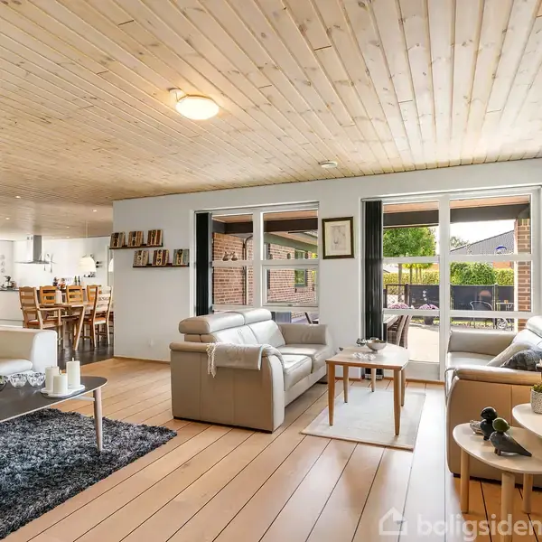 Stue med lyse trægulve og loft, hvor to sofaer og et sofabord står opstillet. I baggrunden ses et spisebord ved siden af åbent køkken. Store vinduer viser terrasse og have.