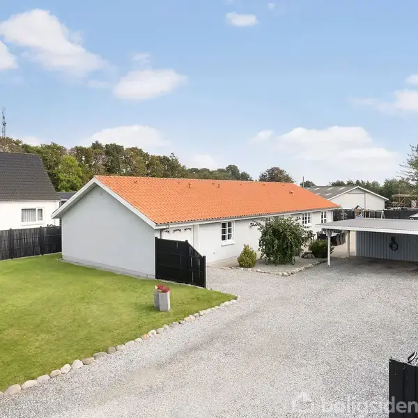 Et hvidt hus med rødt tag står stille ved siden af en carport. Omgivet af en velplejet have og grusbelagt indkørsel, med træer og hegn i baggrunden.