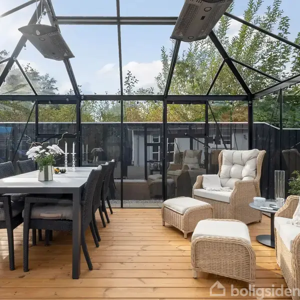 Glashus med spisebord og stole til venstre; loungestole og fodskamler til højre. Omgivet af frodig have, sollys strømmer ind, skaber en lys og indbydende atmosfære.