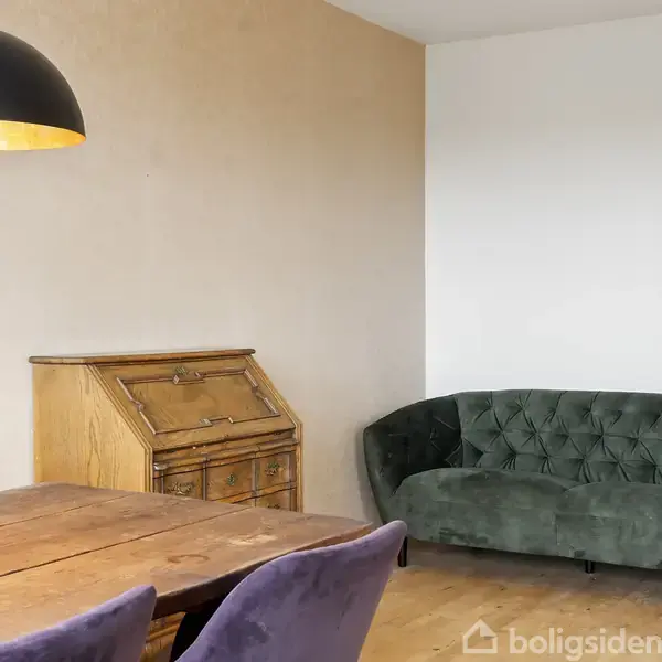 En grøn sofa står i et hjørne ved siden af et antikt skrivebord. En træspisebord med lilla stole og en sort pendellampe i et lyst rum.