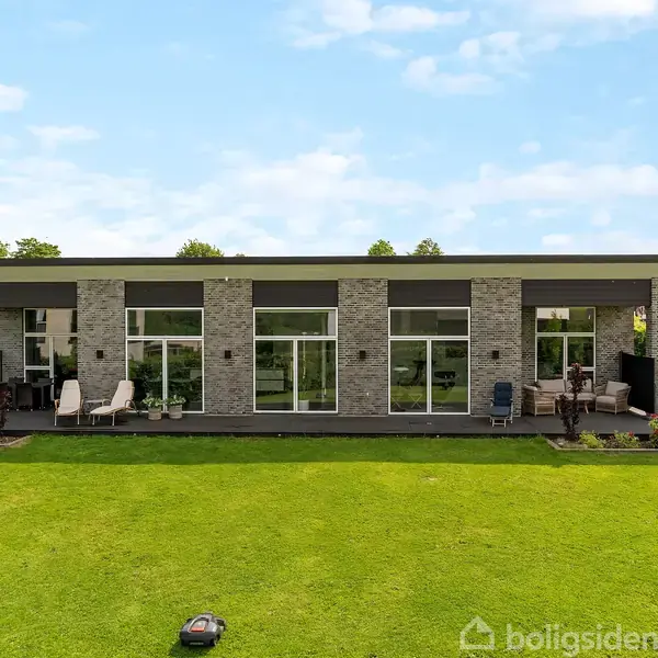 En moderne villa med fladt tag har solpaneler. Foran huset er en anlagt have med en terrasse med bord og liggestole. Omgivet af grønne hække og træer.