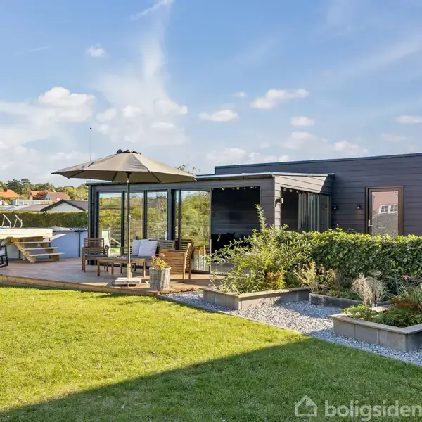 En moderne terrasse med havemøbler og parasol står ved et mørkt facadehus. Græsplæne og planter omringer området, mens en blå himmel giver en solrig stemning.