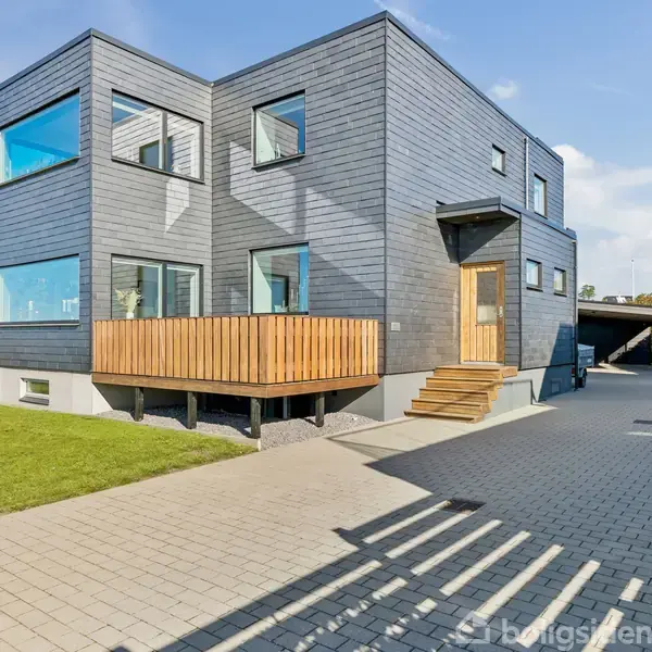 Et moderne hus med fladt tag står stille. Det er bygget i gråt træ og har store vinduer. Omgivet af en lav hæk og beliggende i et villakvarter.