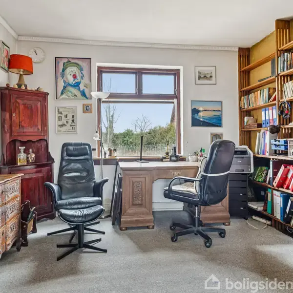Et kontor med to kontorstole ved et træskrivebord. Skabe, kunst på væggene og reoler fyldt med bøger omringer rummet. Vindue giver udsigt til grønne marker.