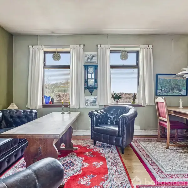 En stue med sorte lædersofaer og et træbord står på røde og hvide tæpper. Store vinduer med gardiner åbner op til lys, mens et spisebord med stole står til højre.