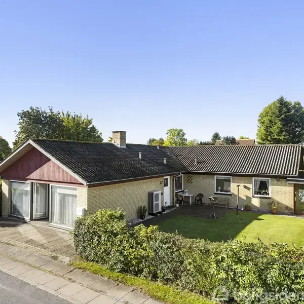 En gulstensvilla med sort tag står ved en vej, omgivet af en velplejet græsplæne og små buske. Der er en lille terrasse med havemøbler foran huset.