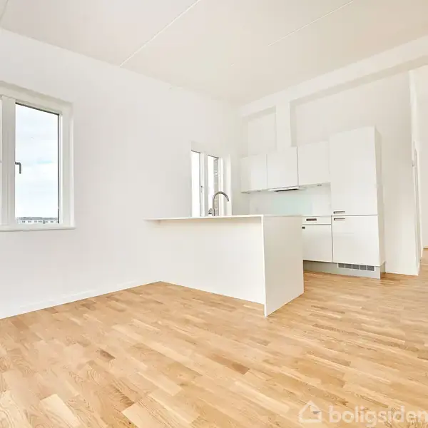 Køkken med hvide skabe og indbygget ovn står i et lyst rum med trægulve. Store vinduer giver udsigt til en moderne bydel.