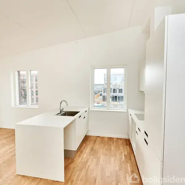 Et moderne køkken med hvide skabe og vask er centreret. Store vinduer lader naturligt lys ind, afslørende en byudsigt udenfor. Trægulv giver en varm kontrast til det hvide interiør.