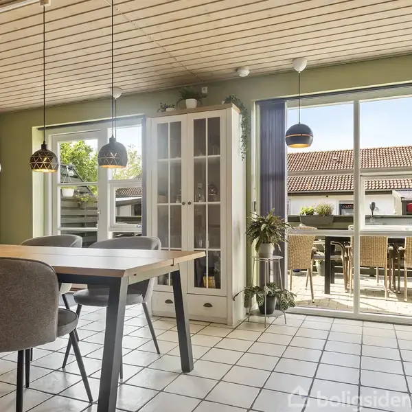 En spisebord med planter og stole står i et køkken med et grønt farvetema, flisegulv og en trappe. Køleskab og ophængte lamper udgør dele af indretningen.