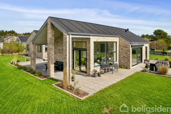 En moderne villa med store vinduer står på en velplejet græsplæne. Den har en fliseterrasse med udendørs møbler. Omgivet af grønne områder og buske under en blå himmel.